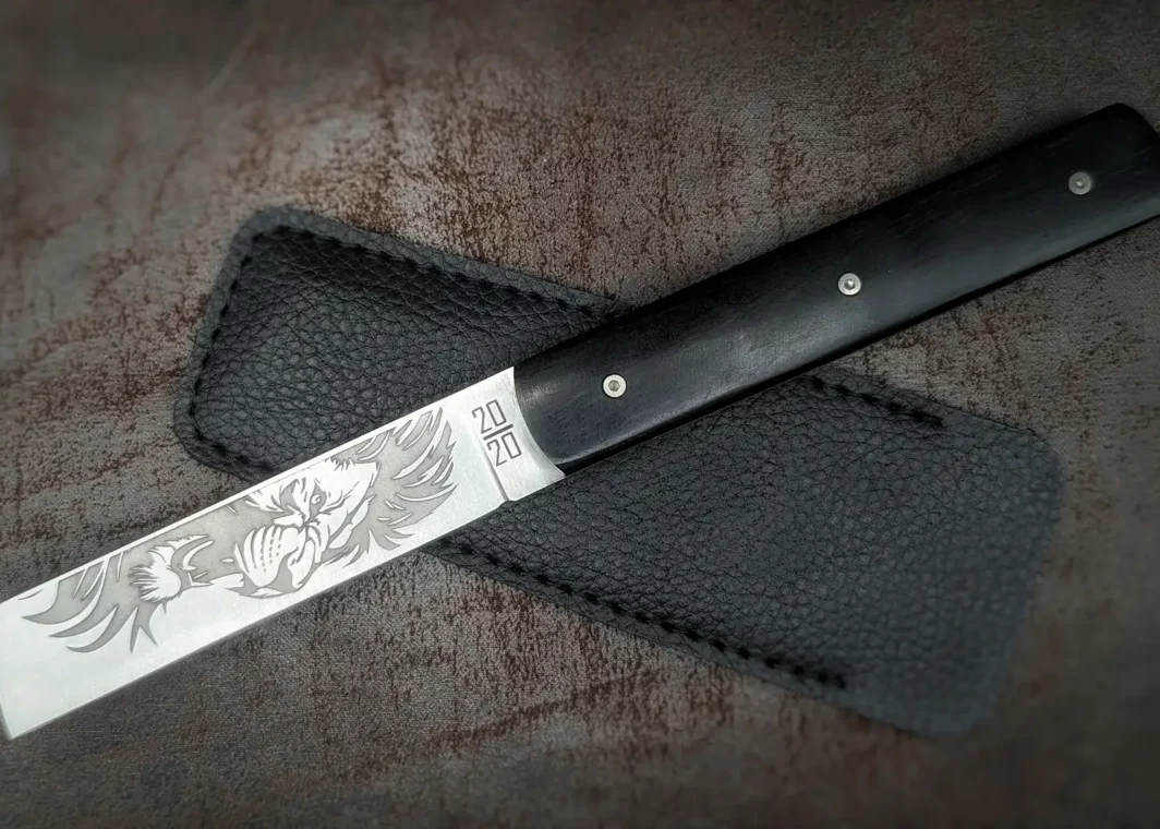 Couteau pliant 20/20 de Le Fidèle. Position ouverte, côté gauche. Le poinçon est visible sur la lame ainsi qu'un décor de lion. Le manche est en ébène. L'étui en cuir est derrière Couteau industriel de marque française Coutellerie Châteauneuf-du-Pape, Coutellerie du Pape, Vaucluse