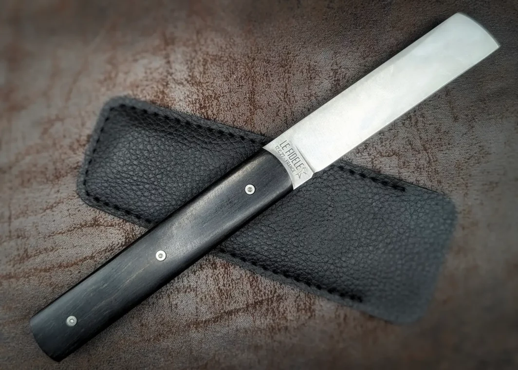 Couteau pliant 20/20 de Le Fidèle. Position ouverte, côté droit. Le manche est en ébène. L'étui en cuir est derrière. Couteau industriel de marque française Coutellerie Châteauneuf-du-Pape, Coutellerie du Pape, Vaucluse