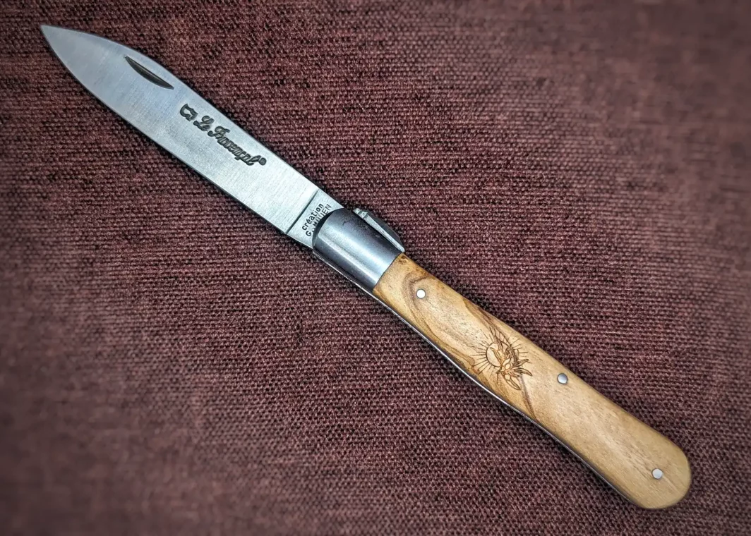 Couteau pliant le Provençal de Au Sabot. Position ouverte, côté gauche. On voit le poinçon sur la lame. Le manche est en olivier avec au centre une petite gravure représentant un soleil et une branche d'olivier. Mitre avant en inox Couteau industriel de marque française Coutellerie Châteauneuf-du-Pape, Coutellerie du Pape, Vaucluse