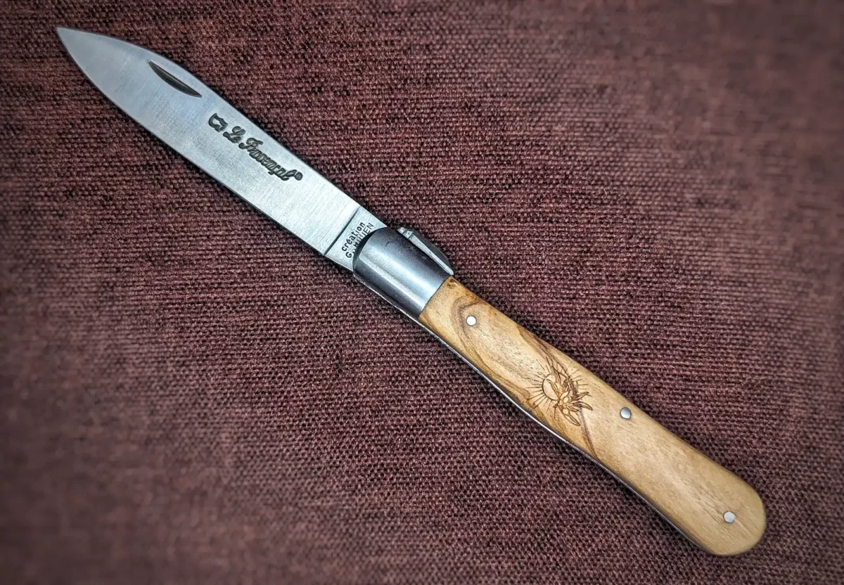 Couteau pliant le Provençal de Au Sabot. Position ouverte, côté gauche. On voit le poinçon sur la lame. Le manche est en olivier avec au centre une petite gravure représentant un soleil et une branche d'olivier. Mitre avant en inox Couteau industriel de marque française Coutellerie Châteauneuf-du-Pape, Coutellerie du Pape, Vaucluse