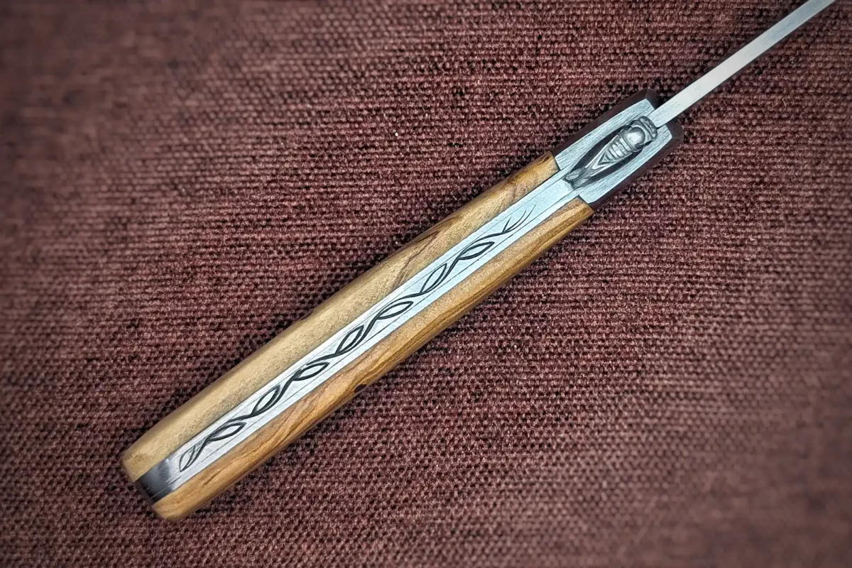 Couteau pliant le Provençal de Au Sabot. Position ouverte, Vue du dos. On voit une guilloche représentant les feuilles d'olivier. Une cigale est placée là où il y a la louche habituellement. Le manche est en olivier. Mitre avant en inox Couteau industriel de marque française Coutellerie Châteauneuf-du-Pape, Coutellerie du Pape, Vaucluse
