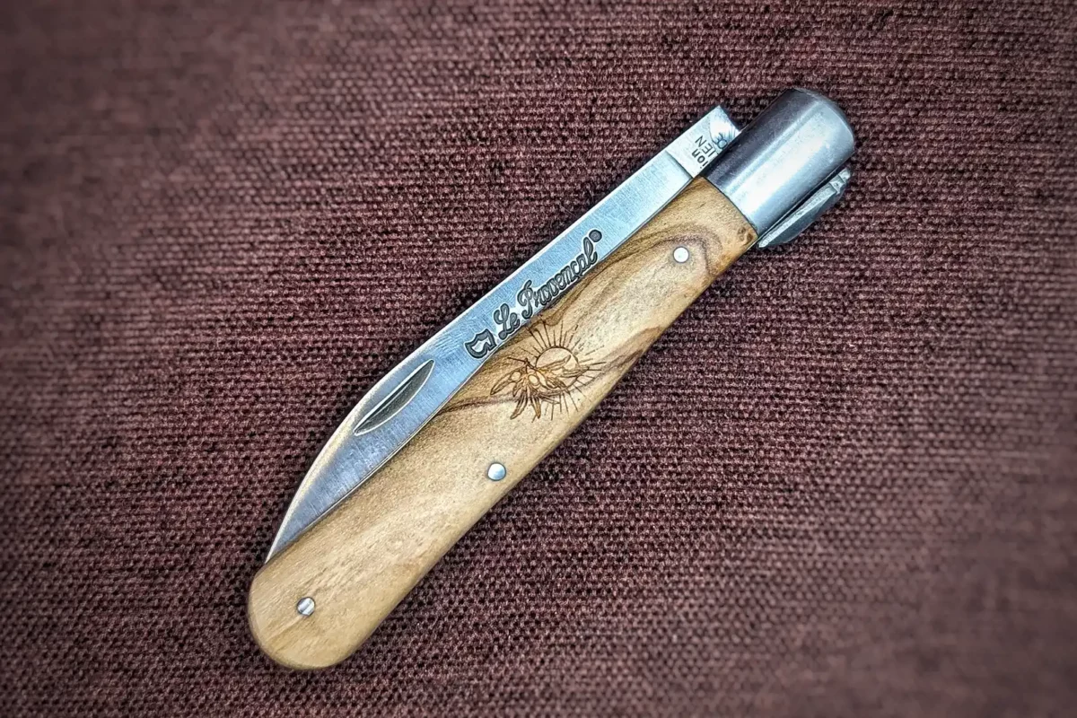 Couteau pliant le Provençal de Au Sabot. Position fermée, côté gauche. On voit le poinçon sur la lame. Le manche est en olivier avec au centre une petite gravure représentant un soleil et une branche d'olivier. Mitre avant en inox Couteau industriel de marque française Coutellerie Châteauneuf-du-Pape, Coutellerie du Pape, Vaucluse