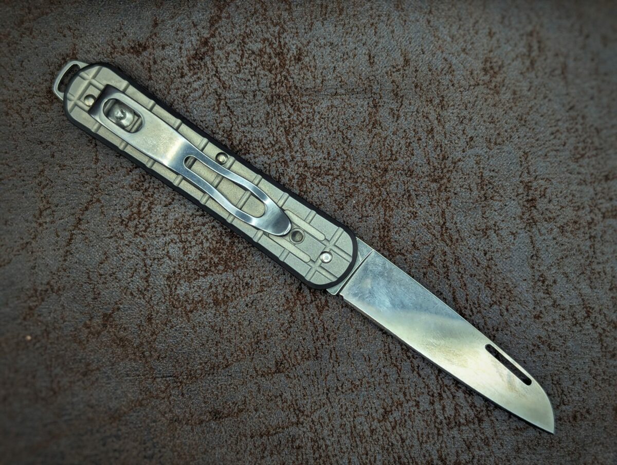Couteau Vulpis, position ouverte, côté droit. Le manche est en titane sablé motif grenade avec un trou pour une lanière. On aperçoit le clip d'attache pour ceinture ou lanière. Couteau industriel de marque italienne Coutellerie Châteauneuf-du-Pape, Coutellerie du Pape, Vaucluse