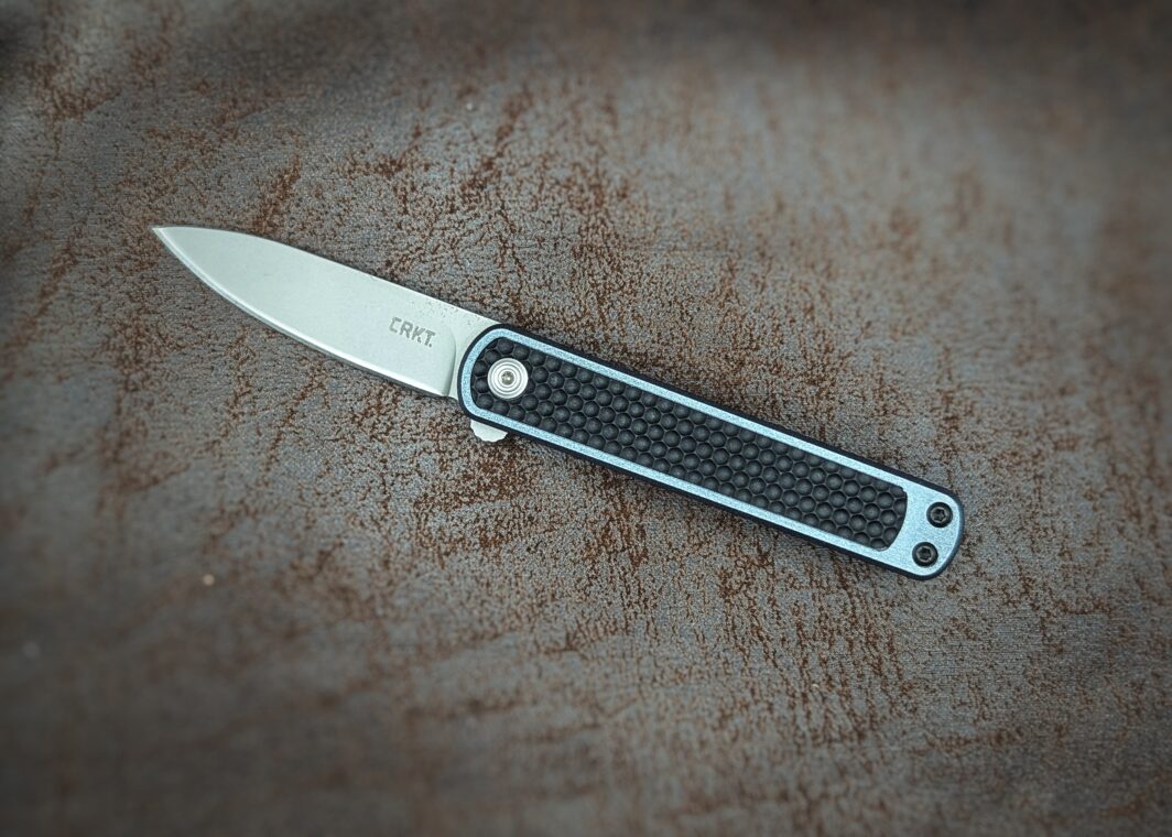 Couteau pliant de CRKT. Position ouverte, côté gauche. Le manche est noir avec un liseré bleu tout autour. On voit la marque écrit sur la lame. Couteau industriel de marque américaine Coutellerie Châteauneuf-du-Pape, Coutellerie du Pape, Vaucluse
