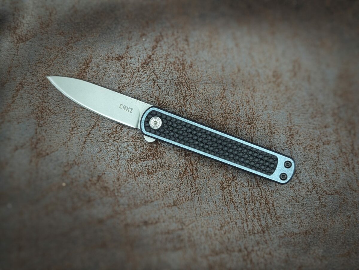 Couteau pliant de CRKT. Position ouverte, côté gauche. Le manche est noir avec un liseré bleu tout autour. On voit la marque écrit sur la lame. Couteau industriel de marque américaine Coutellerie Châteauneuf-du-Pape, Coutellerie du Pape, Vaucluse