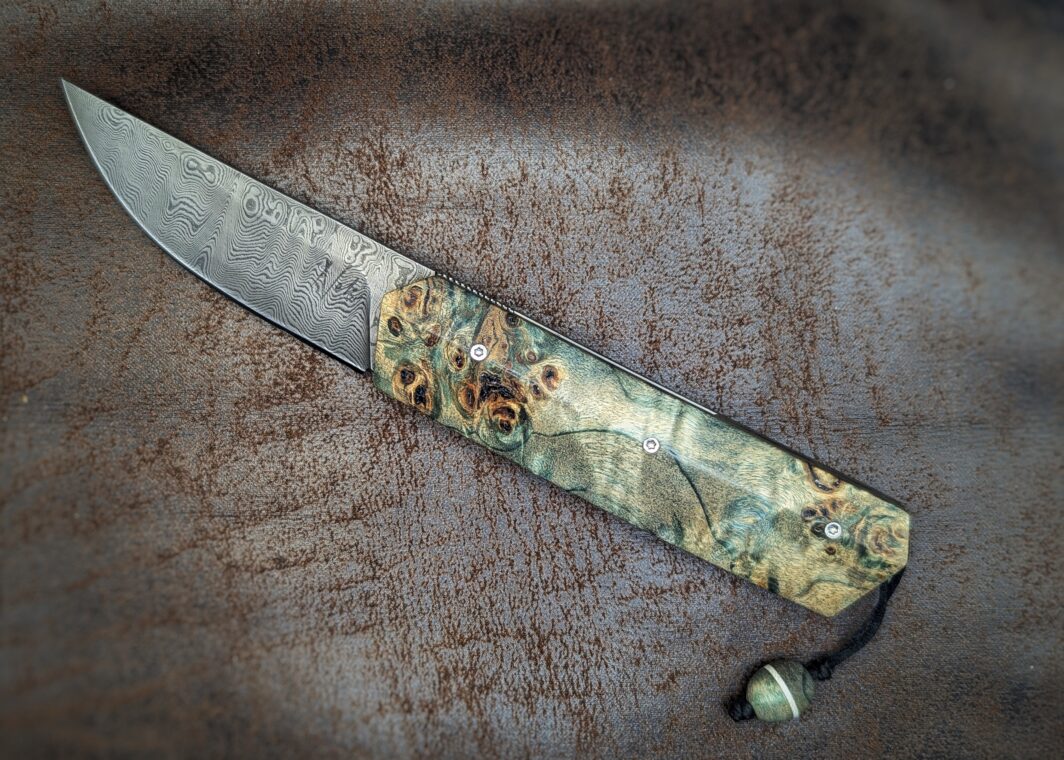 Couteau pliant Liner Lock 'Kiru' de l'artisan Noki. Position ouvert, côté gauche. Lame en damas 208 couches. Manche en Loupe de peuplier stabilisée teintée vert. On voit le poinçon de l'artisan sur la lame. Avec étui en cuir noir. Couteau artisanal français à Châteauneuf-du-Pape, Vaucluse Coutellerie du Pape