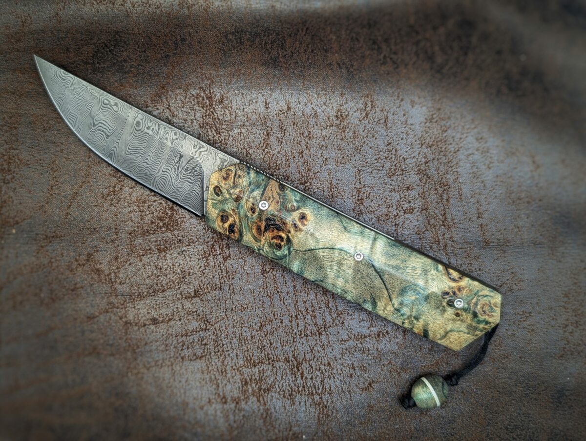 Couteau pliant Liner Lock 'Kiru' de l'artisan Noki. Position ouvert, côté gauche. Lame en damas 208 couches. Manche en Loupe de peuplier stabilisée teintée vert. On voit le poinçon de l'artisan sur la lame. Avec étui en cuir noir. Couteau artisanal français à Châteauneuf-du-Pape, Vaucluse Coutellerie du Pape