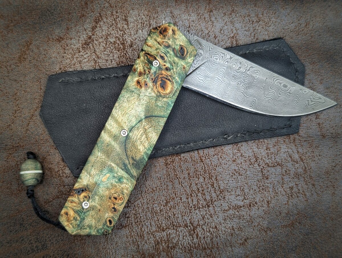Couteau pliant Liner Lock 'Kiru' de l'artisan Noki. Position à demi ouvert, côté droit. Lame en damas 208 couches. Manche en Loupe de peuplier stabilisée teintée vert. Avec étui en cuir noir. Couteau artisanal français à Châteauneuf-du-Pape, Vaucluse Coutellerie du Pape