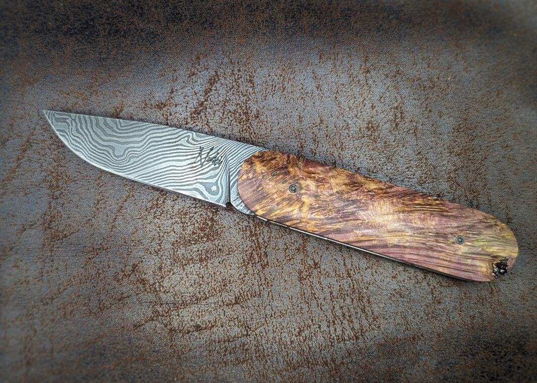 Couteau Le Baldo par l'artisan Noki. Pliant LinerLock FrontFlip. Position ouvert, côté gauche. On voit le poinçon de l'artisan sur la lame damas. Le manche est en bouleau madré stabilisé et teinté violet. Couteau artisanal français à Châteauneuf-du-Pape, Vaucluse Coutellerie du Pape