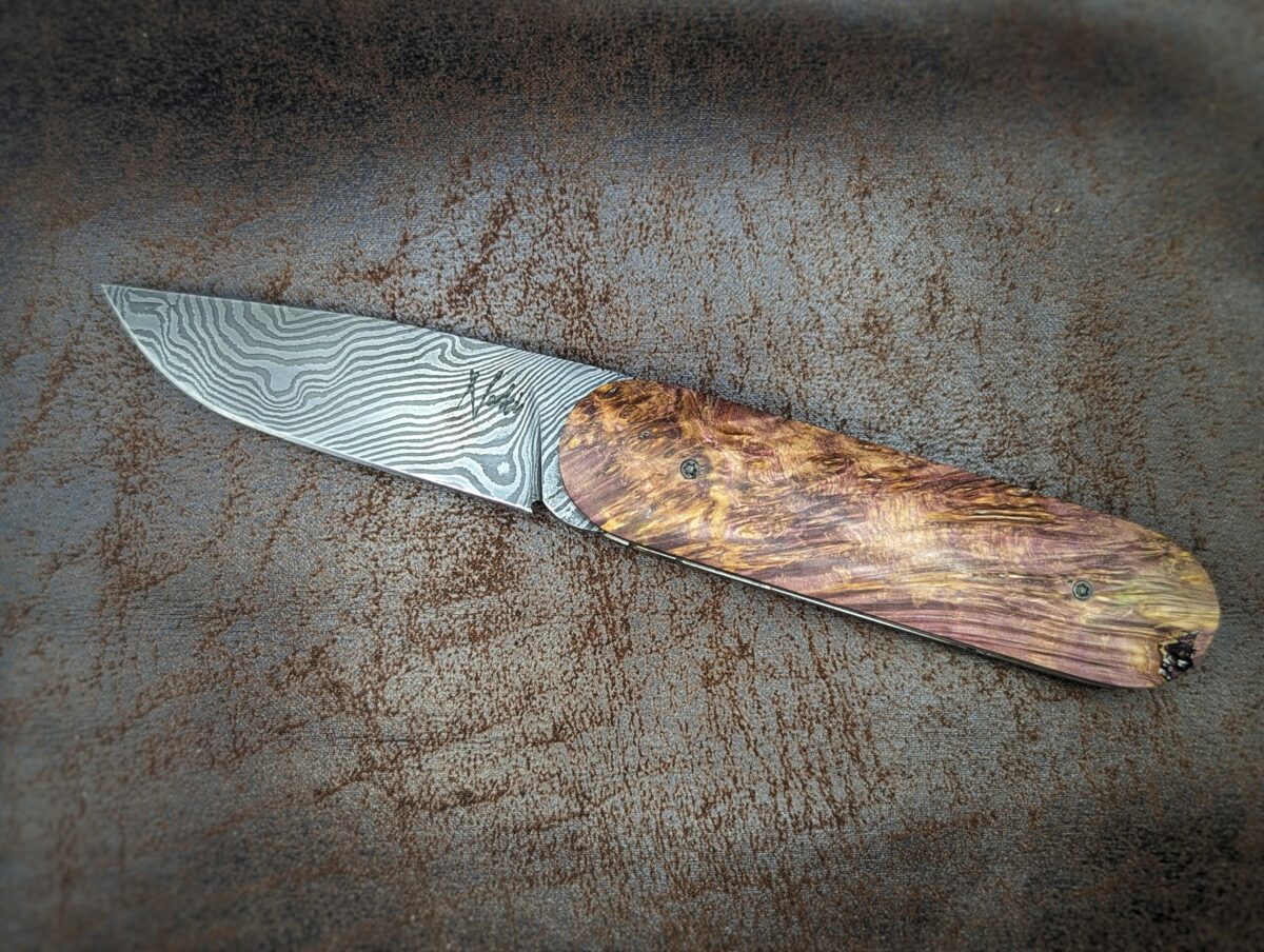 Couteau Le Baldo par l'artisan Noki. Pliant LinerLock FrontFlip. Position ouvert, côté gauche. On voit le poinçon de l'artisan sur la lame damas. Le manche est en bouleau madré stabilisé et teinté violet. Couteau artisanal français à Châteauneuf-du-Pape, Vaucluse Coutellerie du Pape