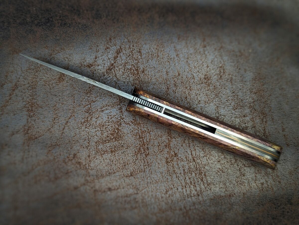 Couteau Le Baldo par l'artisan Noki. Pliant LinerLock FrontFlip. Position ouvert, vue du dessus. La lame est en damas. Le manche est en bouleau madré stabilisé et teinté violet. Couteau artisanal, coutellerie français à Châteauneuf-du-Pape, Vaucluse Coutellerie du Pape