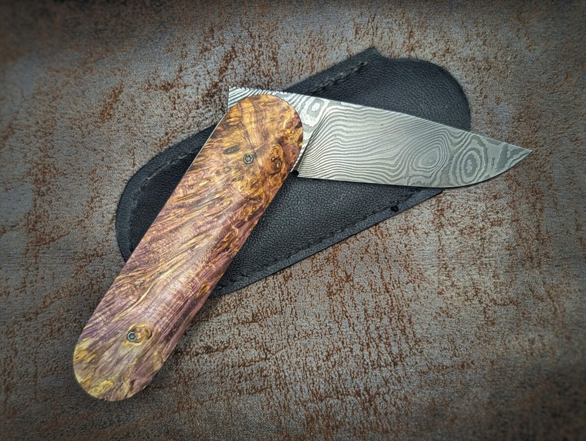 Couteau Le Baldo par l'artisan Noki. Pliant LinerLock FrontFlip. Position demi-ouvert, côté droit. La lame est en damas. Le manche est en bouleau madré stabilisé et teinté violet. On voit l'étui en cuir noir fait main derrière le couteau. Couteau artisanal français à Châteauneuf-du-Pape, Vaucluse Coutellerie du Pape