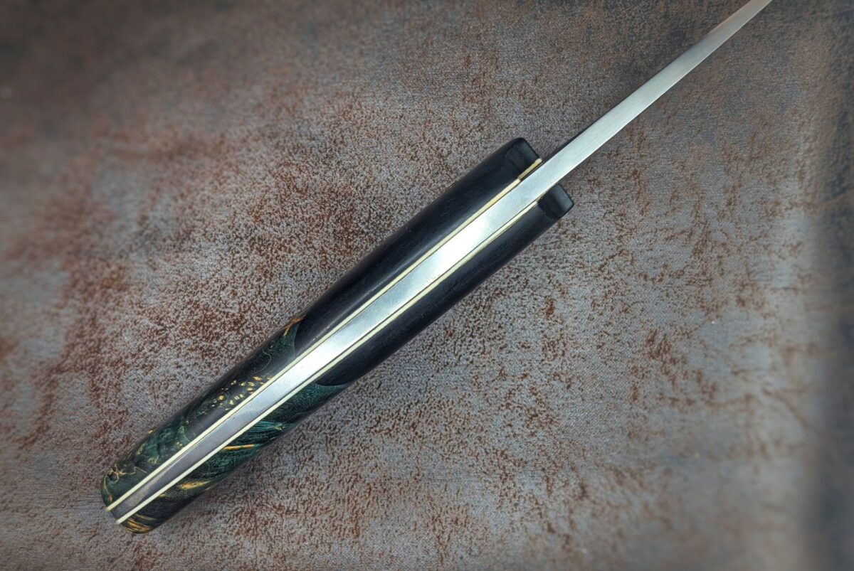 Couteau fixe de l'artisan Lou Marius. Vue du dessus. On voit les intercalaires blancs sur lesquels sont collés les matériaux du manche. Le manche s'affine vers l'arrière du couteau. Couteau artisanal, coutellerie français à Châteauneuf-du-Pape, Vaucluse Coutellerie du Pape