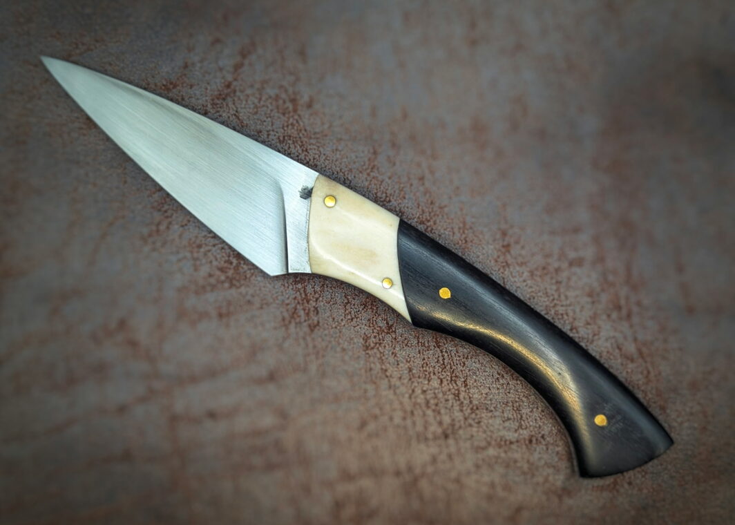 Couteau fixe par l'artisan Dahéna Urli. Vue côté gauche. Sa lame est en acier carbone C130 avec finition tirée en long. Son manche est en ébène avec mitre en bois de renne. Couteau artisanal, coutellerie français à Châteauneuf-du-Pape, Vaucluse Coutellerie du Pape