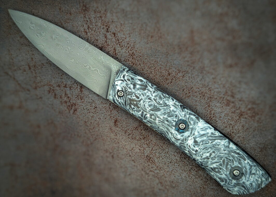 Couteau Piémontais Full Carbone par l'artisan Dahéna Urli. Position ouverte, côté gauche. Le manche est en fibre de carbone marbrée noir et blanc. Au niveau des vis, un insert rond en fibre de carbone paillette bleu. La lame est en damas carbone. Couteau artisanal, coutellerie français à Châteauneuf-du-Pape, Vaucluse Coutellerie du Pape