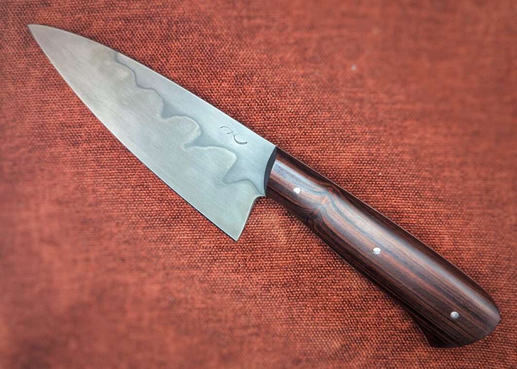 Couteau fixe de cuisine par l'artisan Alban Campagnola. Vue du côté gauche. La lame est en XC100 avec une révelation d'une trempe sélective au perchlorure de fer. On voit sur la lame le poinçon d'Alban. Le manche est en Palissandre de santos. Couteau artisanal, coutellerie français à Châteauneuf-du-Pape, Vaucluse Coutellerie du Pape
