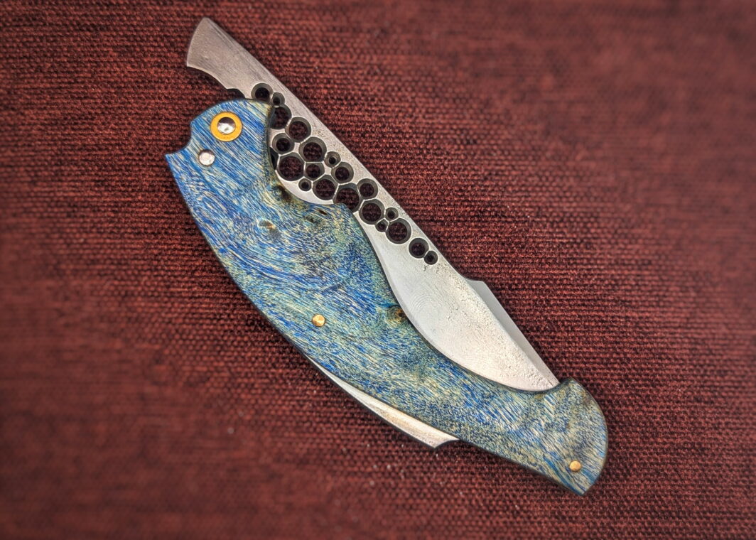 Couteau pliant Amphiptère de l'artisan Dracoforge. Position fermé, côté droit. La lame est en Z40 et le manche en Peuplier stabilisé bleu. La fausse pièce dépassant du dessus rappel un univers marin. Couteau artisanal, coutellerie français à Châteauneuf-du-Pape, Vaucluse Coutellerie du Pape