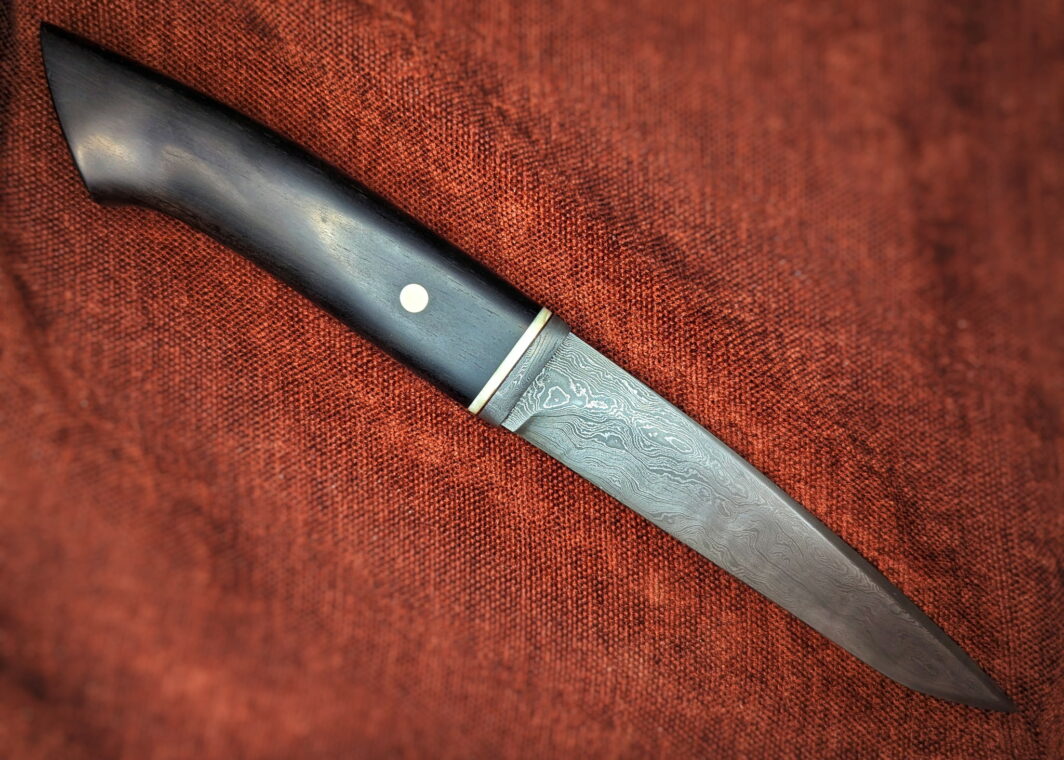 Couteau fixe artisanal par l'artisan Jean-Paul Sire. Vue du côté droit. La lame est en acier damassé 300 couches forgé. Le manche est en ébène avec bague en ivoire de phacochère et damas. Il y a un insert en ivoire de phacochère sur le manche. Couteau artisanal, coutellerie français à Châteauneuf-du-Pape, Vaucluse Coutellerie du Pape