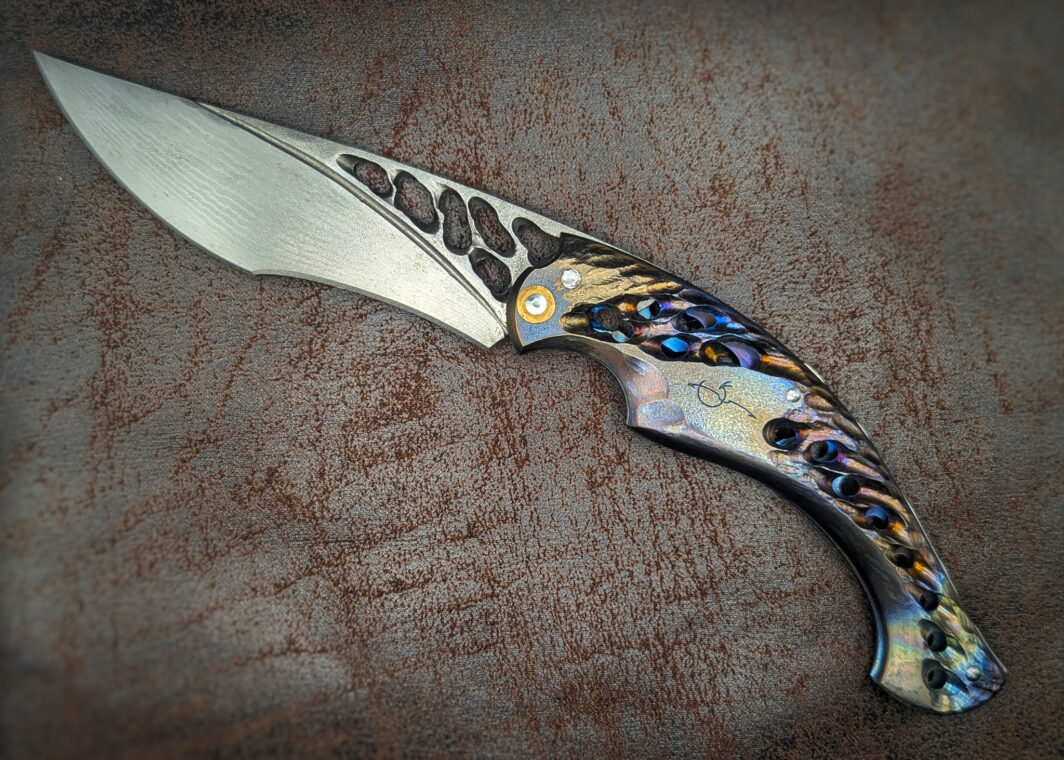 Couteau Neptune 56 par l'artisan Dracoforge. Position ouvert. Côté gauche. Le manche est en titane coloré et ajouré. La lame est en Suminagashi VG-10 ajourée également. Couteau artisanal, coutellerie français à Châteauneuf-du-Pape, Vaucluse Coutellerie du Pape