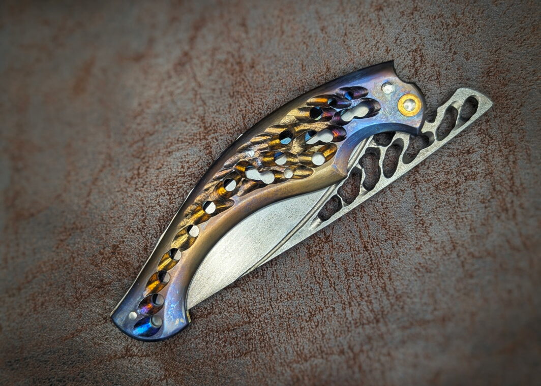 Couteau Neptune 56 par l'artisan Dracoforge. Position fermée. Côté droit. Le manche est en titane coloré et ajouré. La lame est en Suminagashi VG-10 ajourée également. Couteau artisanal, coutellerie français à Châteauneuf-du-Pape, Vaucluse Coutellerie du Pape