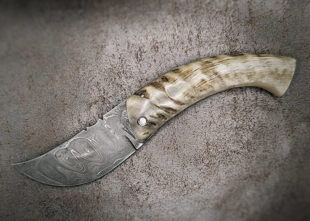 couteau pliant deux clous avec manche en corne de bélier et lame en damas carbone position ouverte, coté gauche