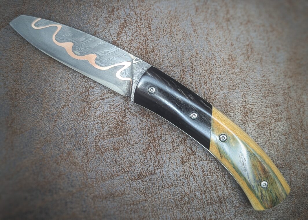 Couteau pliant artisanal par Dahéna Urli. Lame en damas cuivre manche en ivoire de mammouth et corne de buffle. Position ouvert, côté gauche