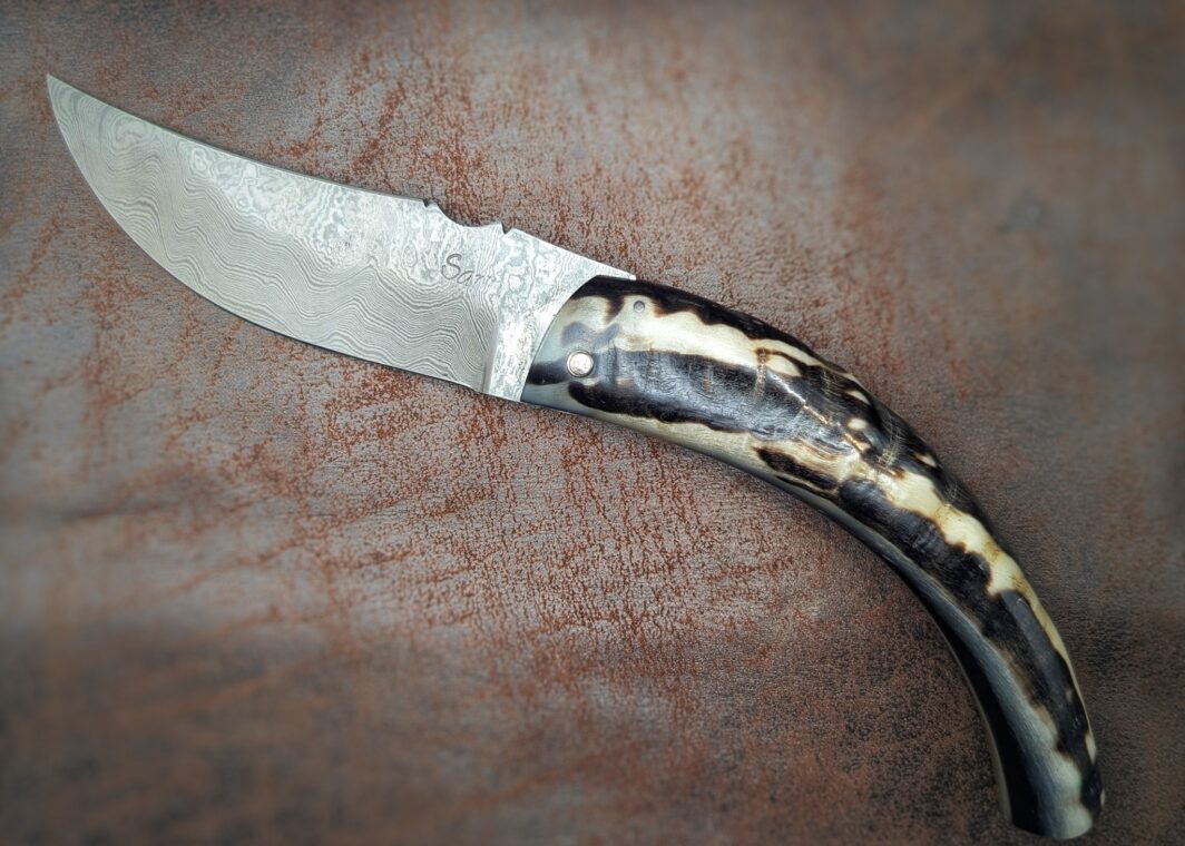 Couteau pliant artisanal par José Viale. Lame en damas carbone, manche en corne de bélier noire et blanche. Position ouverte, vue du côté gauche