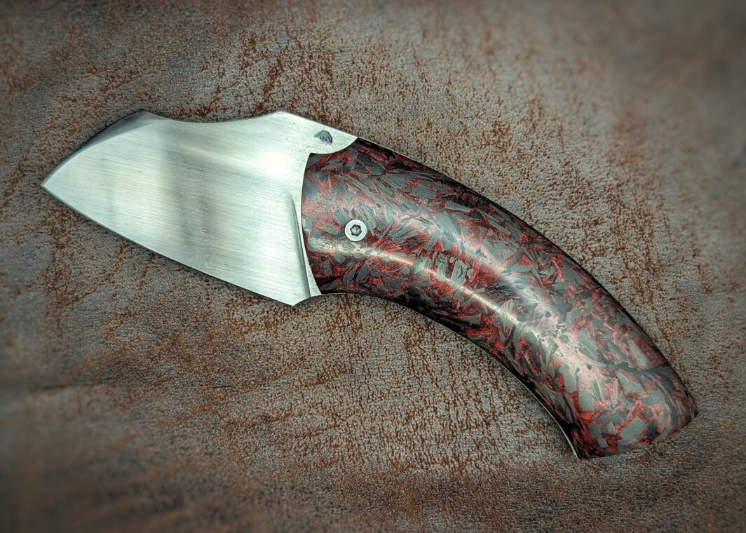 Couteau pliant artisanal par Lou Marius. Lame en acier carbone C130. Manche en fibre de carbone paillette rouge. Position ouverte, vue du côté gauche.