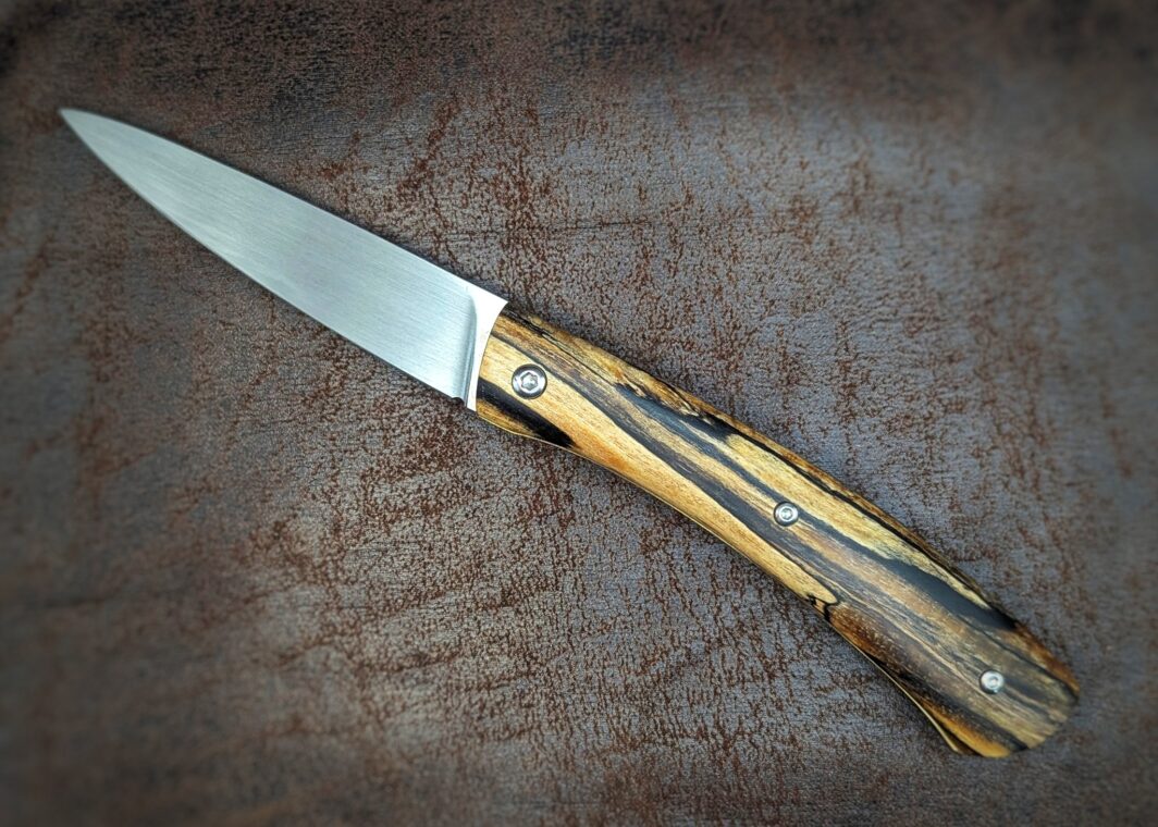 Couteau cran forcé artisanal par Dahéna URLI. Lame en inox Z40 et manche en hêtre. Platines en laiton avec guillochage. Position ouvert, côté gauche